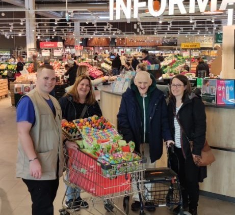 Das Foto zeigt drei Personen in einem Raum, die alle ein Lächeln im Gesicht haben. In der Mitte des Bildes steht Herr Schiller von Globus, der eine Tüte mit Schokoladeneiern in der Hand hält. Links von ihm steht die Leitung des Wohnverbunds Eichborn, die ebenfalls ein Lächeln im Gesicht hat. Rechts von Herrn Schiller steht ein Bewohner des Wohnverbunds, der sich freut, dass er eine Tüte mit Schokoladeneiern geschenkt bekommen hat.